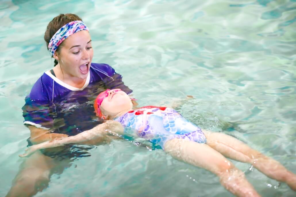 Dolphin Swim Club instructor helping a smiling young swimmer float on her back