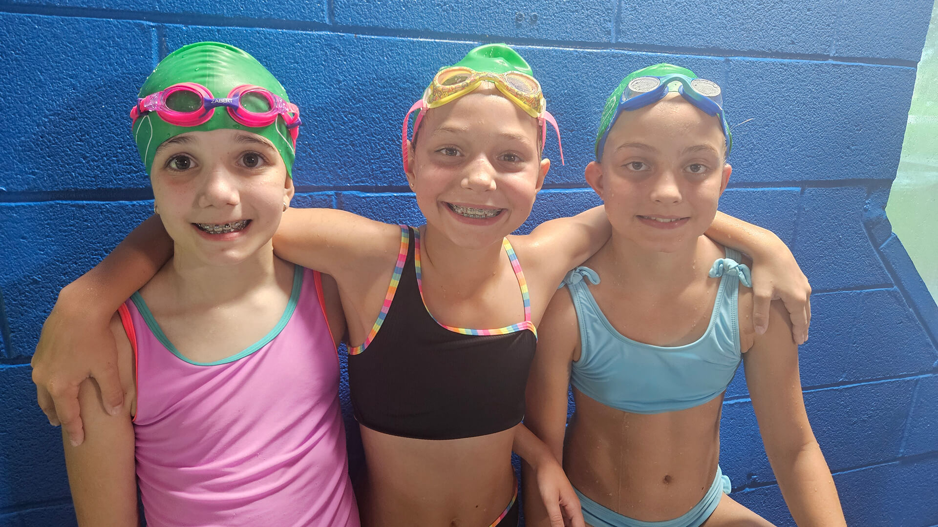 Three Dolphin Swim Club swim team girls smiling in goggles and caps